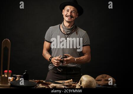 Portrait of handsome Leather goods craftsman in stylish hat doing handbags and leather accessories for ladies in his workshop on black dark background Stock Photo