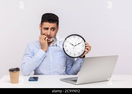 Deadline concept. Nervous anxious man employee sitting in office workplace holding big clock, biting finger nails and looking worried about time limit Stock Photo