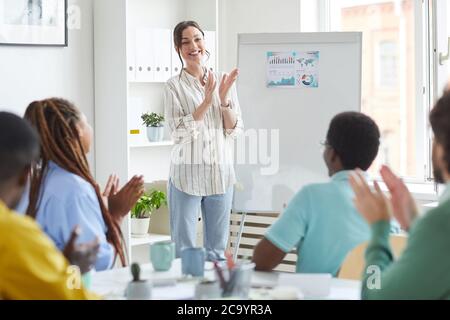 Portrait of smiling female leader applauding while standing by whiteboard in conference room and congratulating multi-ethnic business team on successful launch, copy space Stock Photo