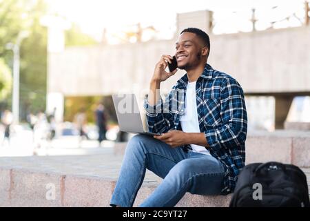 Black man using laptop, sitting in the city with smartphone Stock Photo