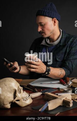 Cobbler Man using smart phone and holding cup of coffee in front of table with tools at shoemaking workshop Stock Photo