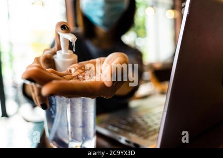 Young woman washing hands with alcohol gel in quarantine for coronavirus wearing protective mask with social distancing and using laptop computer work