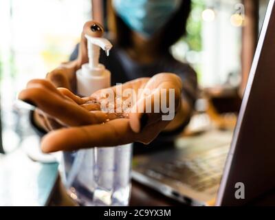 Young woman washing hands with alcohol gel in quarantine for coronavirus wearing protective mask with social distancing and using laptop computer work
