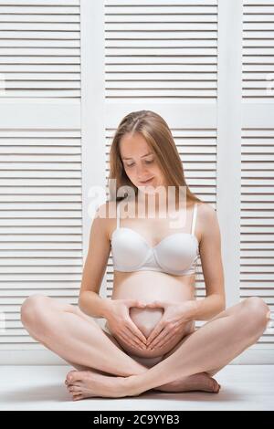 Young beautiful pregnant woman sitting in pose of Lotus and put hands in heart shape on stomach. Vertical frame. Stock Photo