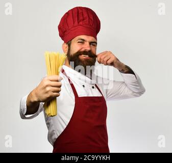 Man with beard isolated on white background. Chef holds bunch of spaghetti in hand. Catering and italian food concept. Cook with happy face in burgundy uniform holds dry pasta curling his moustache. Stock Photo