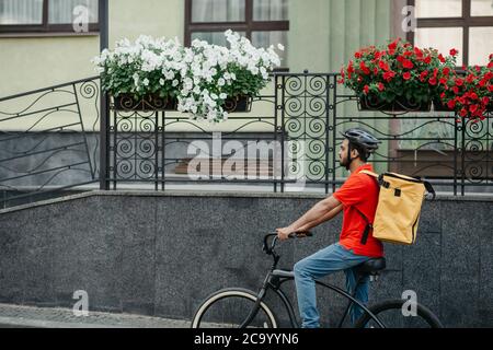 Courier carry out orders for delivery of goods. Man in helmet and big yellow backpack rides bicycle