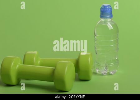 Dumbbells in bright green color and water bottle isolated on green background. Sports and healthy regime equipment. Bottle near lightweight barbells, copy space. Athletics and weight loss concept. Stock Photo