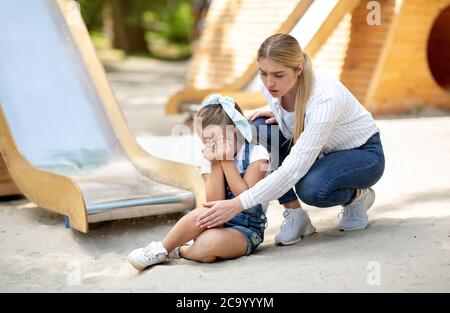 Mother Examining Daughter's Injured Knee After Slide Ride On Playground Stock Photo