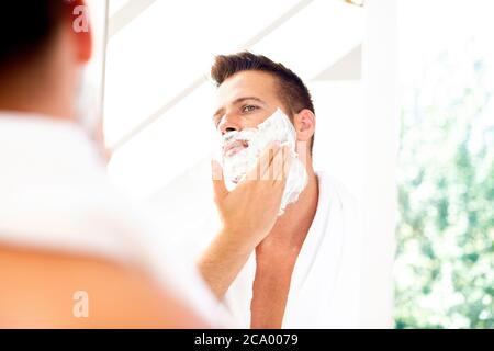Close-up shot of handsome man using shaving foam before shaving while standing at bathroom mirror. Stock Photo