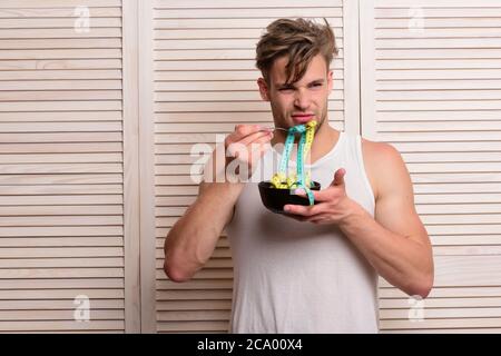 Weight management and diet concept. Guy with disgusted face expression on beige jalousie background. Fitness lifestyle and regime idea. Man with unshaven face holds bowl and fork with measuring tape Stock Photo