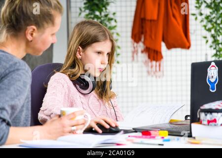 Young, blond girl in pink sweater is confused while doing her lessons. Her mother is helping her. Homeschooling during isolation at home. Stock Photo