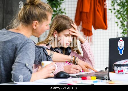 Teenage, blond girl in pink sweater is confused and asking her mother for help while doing her lessons. Mother is drinking a tea and trying to help. Stock Photo