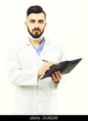 Man with concentrated face in white coat and surgical mask isolated on white background. Doctor with beard writes history of present illness in notebook. Treatment and ambulance services concept Stock Photo