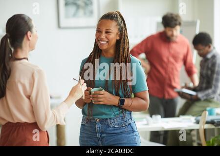 Waist up portrait of contemporary African-American woman smiling cheerfully while talking to female colleague during coffee break in office, copy space Stock Photo