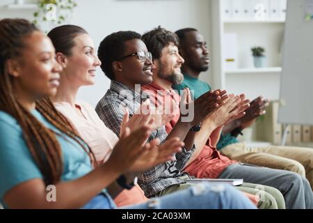 Side view portrait of multi-ethnic group of people applauding while sitting in row in audience or conference room, copy space