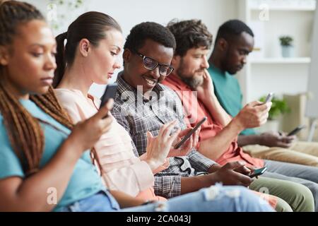 Portrait of multi-ethnic group of people sitting in row and using smartphones while waiting for conference, focus on smiling African-American man talking to female colleague beside him, copy space Stock Photo