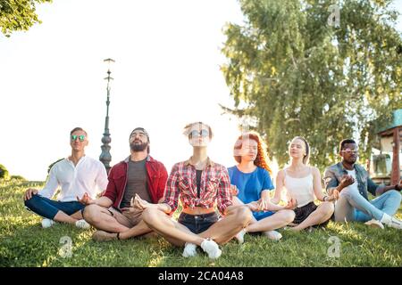Time for yoga, Attractive young multicultural friends exercising in yoga lotus pose while resting after a long trip in Botanical garden. Multiethnic p Stock Photo