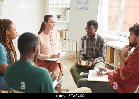 Multi-ethnic group of people sitting in circle while discussing strategy for business project in office, focus on female leader talking to colleagues, copy space Stock Photo