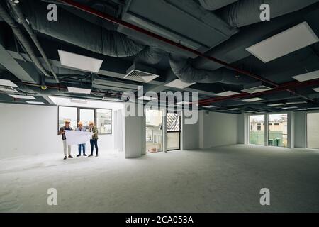 Group of young construction engineers in hardhats checking blueprint of building they are working on Stock Photo