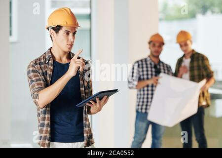 Handsome young civil engineer in hardhat and plaid shirt inspecting construction site and taking notes in document on tablet computer Stock Photo