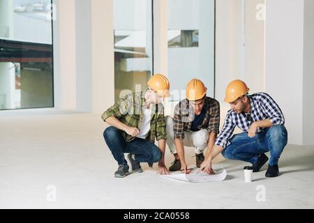 Serious young construction engineers in hardhats discussing construction plan on the floor in empty room Stock Photo