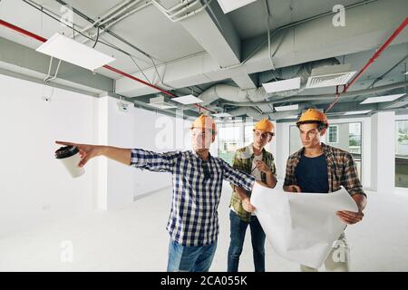 Contractor with cup of coffee in his hand pointing at pipes on the wall and showing it to colleagues with blueprint Stock Photo