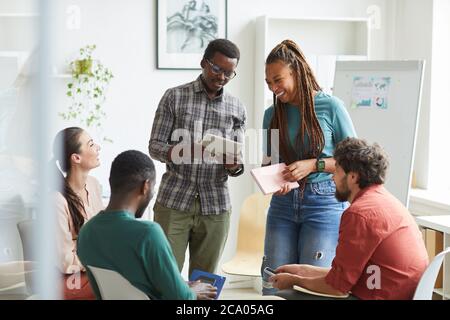 Multi-ethnic group of people sitting in circle while discussing business project in office, focus on smiling African-American woman talking to colleague standing up , copy space Stock Photo