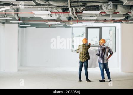 Young construction engineers in plaid shirts and hardhats holding blueprint and looking at city through window Stock Photo