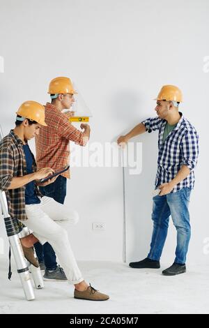 Construction workers measuring wall with various tool when their colleague enterring data in document on tablet computer Stock Photo