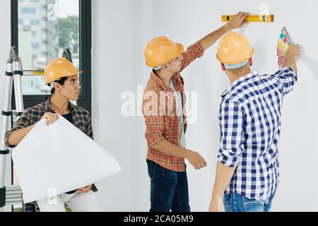Construction workers discussing how to pain walls in empty room, their colleague checking blueprint Stock Photo