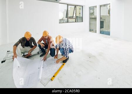 Team of young builders in hard hats checking blueprints before starting renovation process in apartment of client Stock Photo