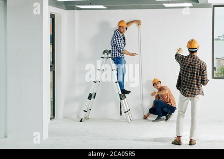 Team of construction workers in hardhats measuring wall with tape measure Stock Photo