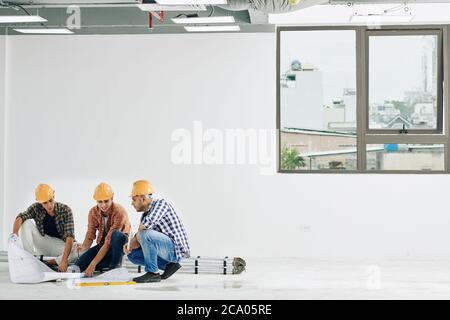 Team of construction workers checking blueprints of the floor of empty room in industrial building Stock Photo