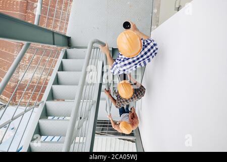 Construction workers in hard hats walking up the stairs one after another, view from the top Stock Photo