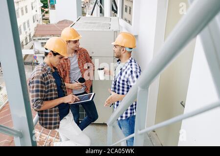 Young construction workers standing on balcony, drinking coffee, discussing news and checking plan of works during break Stock Photo