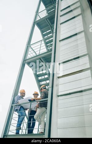 Team of young construction engineers standing on stairs and discussing details in blueprint and planning works Stock Photo