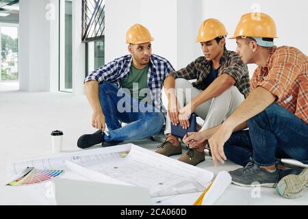 Young serious builders in hardhats sitting on the floor and discussing how to renovate apartment Stock Photo