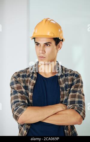 Portrait of handsome serious young mixed-race young builder in hardhat crossing arms and looking away Stock Photo