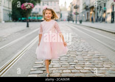 Cute blond little girl walks along the street of the old European city in her beautiful pink dress. Front view. Copy space