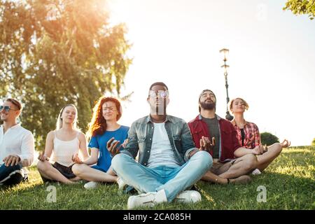 Time for yoga, Attractive young multicultural friends exercising in yoga lotus pose while resting after a long trip in Botanical garden. Multiethnic p Stock Photo
