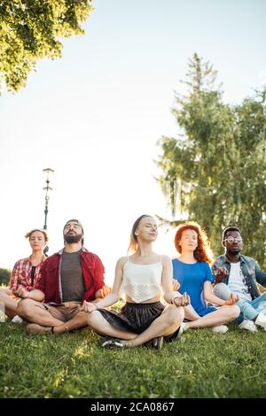 Diverse adult caucasian and afro american friends enjoying relaxing yoga outdoors in the park. Practicing yoga in nature landscape, with blonde woman Stock Photo