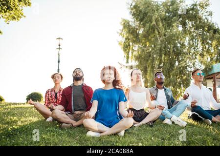 Group of diverse young girls and guys in street casual wear doing physical training with coach on green grass in summer park outdoors. Stock Photo