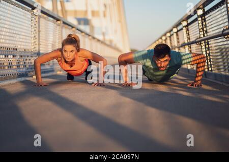 Young couple is exercising outdoor on bridge in the city. They are doing push-ups. Stock Photo