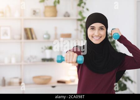 Indoor Sport Concept. Positive Arabic Woman Training With Dumbbells At Home Stock Photo