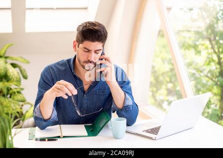 Shot of unhappy businessman making a call and writing something while sitting behind his laptop at office desk. Stock Photo