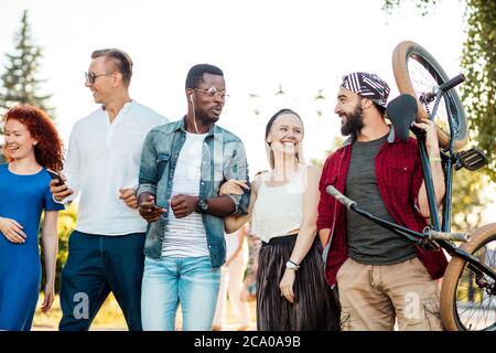 Multi-ethnic group of people going with their male friend to bmx contest in the urban summer park, Young cheerful man giving piggyback ride to his blo Stock Photo