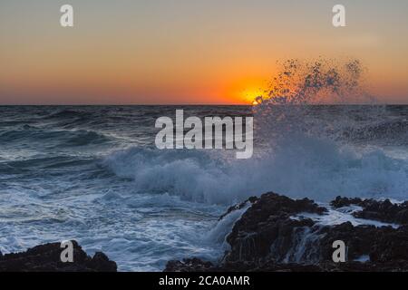 Sunset and storm at sea. Large waves against the background of the setting sun. Summer storm on the Black sea. Beautiful sea spray on the rocks. Stock Photo