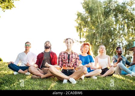 Diverse yoga class of cheerful young friends in summer casual wear sitting in lotus pose on green lown in the public park, front view with focus on be Stock Photo
