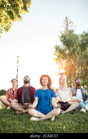 Time for yoga, Attractive young multicultural friends meditating in yoga lotus pose while resting after a long trip in Botanical garden. Multiethnic p Stock Photo
