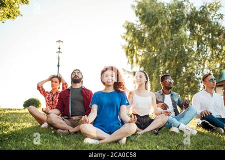 Time for yoga, Attractive young multicultural friends exercising in yoga lotus pose while resting after a long trip in Botanical garden. Multiethnic p Stock Photo